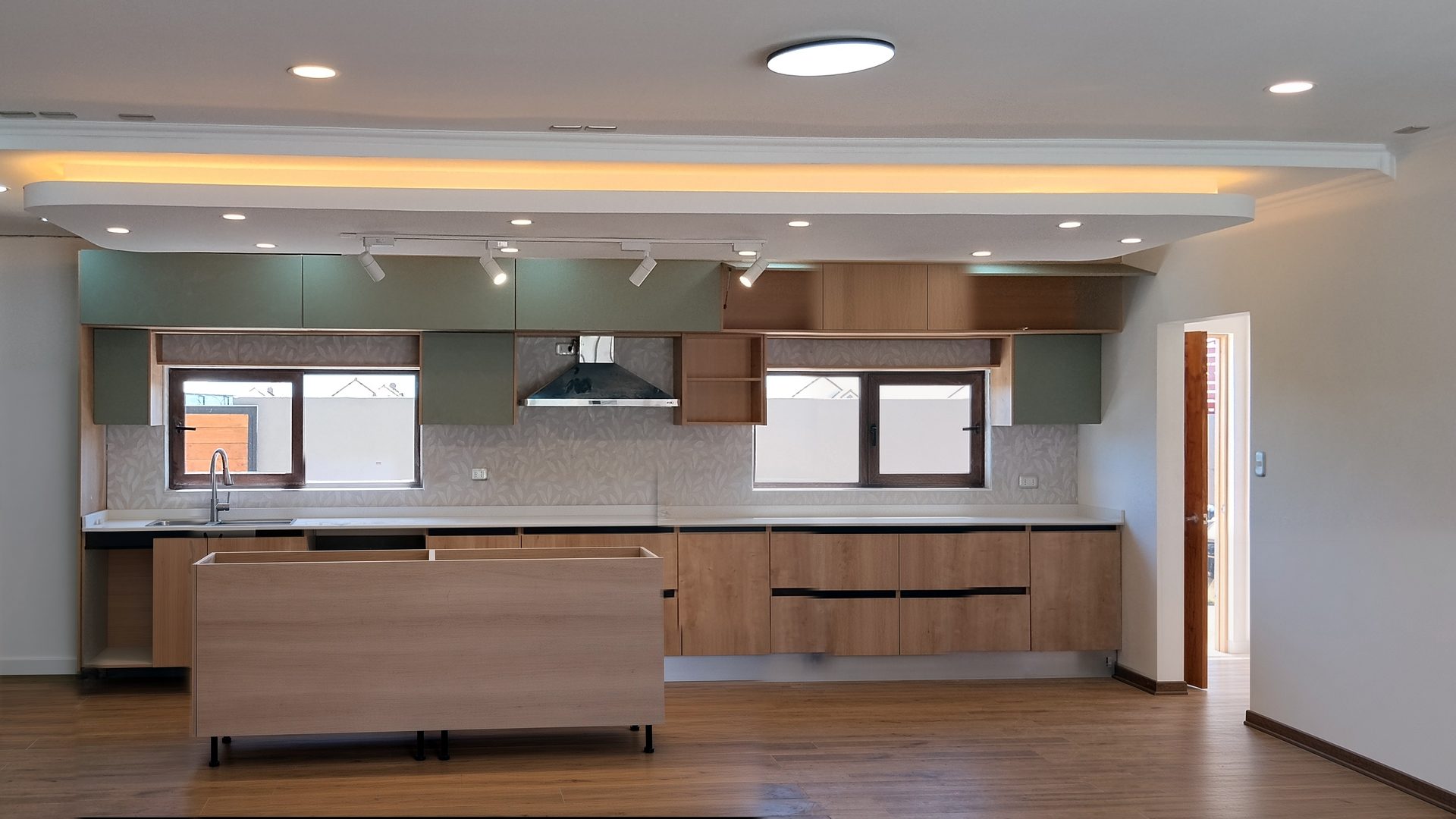 Cocina con cenefa de cielo de esquinas redondeadas, luz LED indirecta cálida, focos embutidos y riel de focos sobre muebles verde salvia y de madera